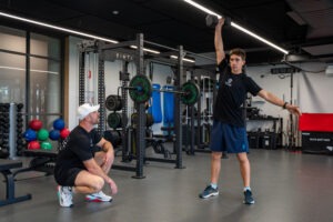 An SCBC student athlete performing a single-arm overhead dumbbell snatch while Coach Michael Rabie crouches to monitor his lifting form in the college gym.