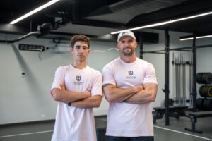 Medium portrait of Coach Andy Price and an SCBC student athlete smiling slightly with arms crossed, wearing matching pastel pink South Coast Baptist College sports shirts in the gym.