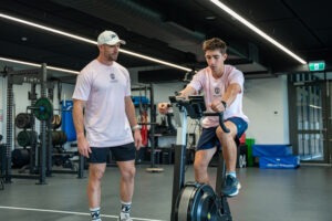 South Coast Baptist College Coach Andy Price monitoring a student athlete's performance and console data on a Concept2 BikeErg in the campus gym.