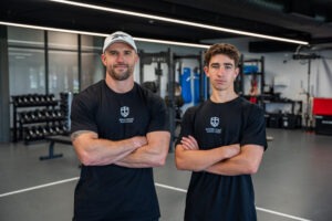Portrait of South Coast Baptist College Strength and Conditioning Specialist Andy Price and a male student athlete standing side-by-side with arms crossed in the High-Performance Centre, wearing matching black SCBC training shirts.